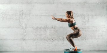 In palestra, con muro bianco, una donna con leggings e top con la coda di cavallo su la bosu ball esegue uno squat correttamente