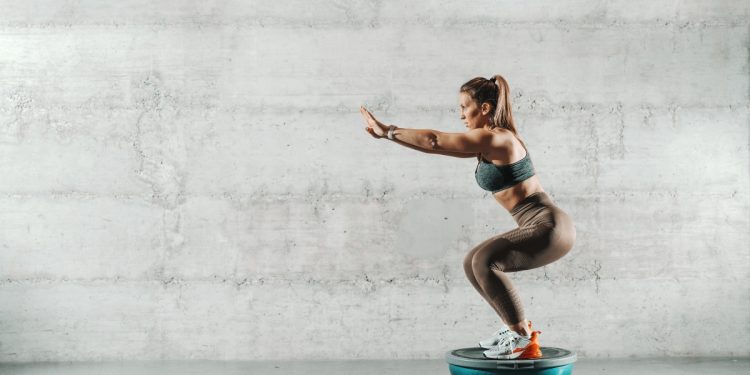 In palestra, con muro bianco, una donna con leggings e top con la coda di cavallo su la bosu ball esegue uno squat correttamente