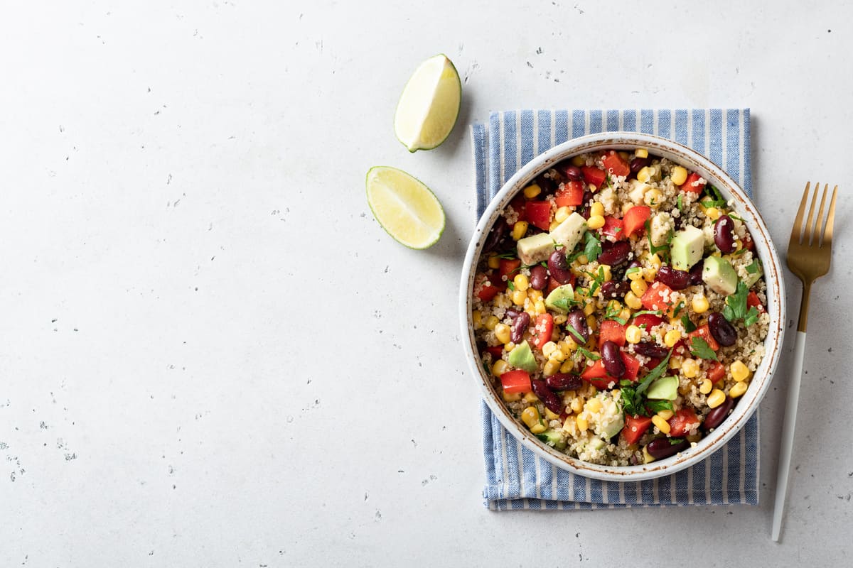 Im primo piano su un tavolo di marmo bianco in un piatto fondo c'è della quinoa con verdure. Sulla destra una forchetta e in alto a sinistra due spicchi di lime.