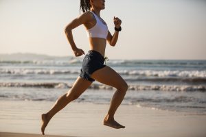 donna che ha scelto come sport anticellulite la corsa. Corre con gli shorts e top bianco sulla spiaggia in riva al mare piedi nudi