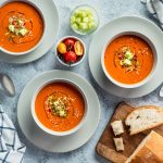 Gazpacho: la ricetta originale della zuppa fredda spagnola 1 su tavolo di marmo, piatti bianchi con 3 ciotole di gazpacho con pezzi di cetrioli, al centro ciotole con pomodorini e un'altra ciotola con cetrioli tagliati a cubetti. Sulla destra tagliere con fette di pane