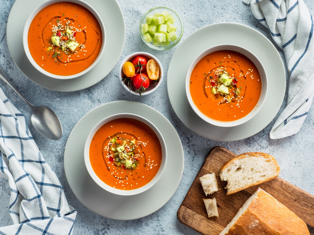 su tavolo di marmo, piatti bianchi con 3 ciotole di gazpacho con pezzi di cetrioli, al centro ciotole con pomodorini e un'altra ciotola con cetrioli tagliati a cubetti. Sulla destra tagliere con fette di pane