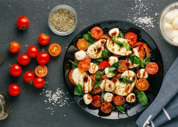 tavolo nero visto dall'alto con un piatto di insalata caprese e con ingredienti, sale, mozzarella, pomodori e origano