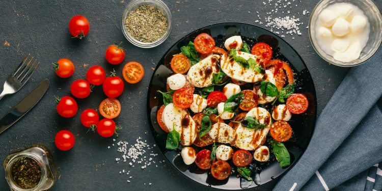 tavolo nero visto dall'alto con un piatto di insalata caprese e con ingredienti, sale, mozzarella, pomodori e origano