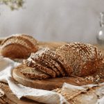 In primo piano, su un tavolo di legno chiaro, su un tagliere c'è del pane integrale tagliato a fettine sottili.