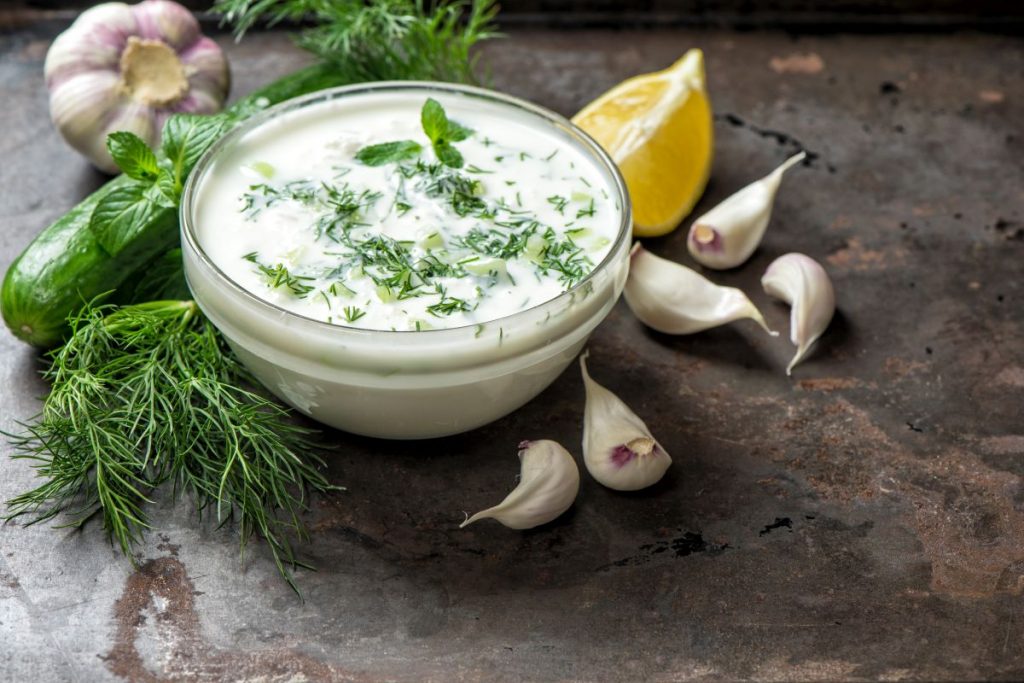 su tavolo da cucina, ciotola di vetro con la salsa allo yogurt, attorno gli ingredienti limone, erbe aromatiche e aglio