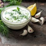 su tavolo da cucina, ciotola di vetro con la salsa allo yogurt, attorno gli ingredienti limone, erbe aromatiche e aglio