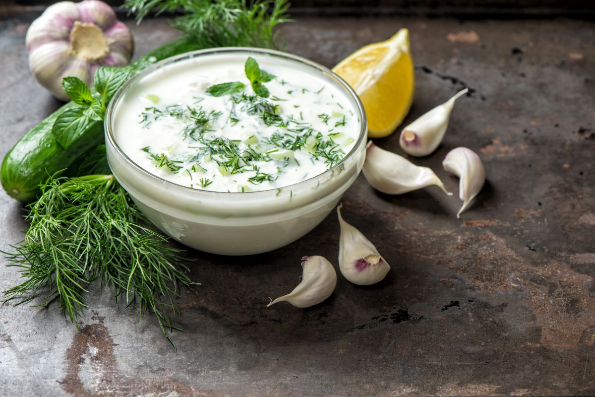 su tavolo da cucina, ciotola di vetro con la salsa allo yogurt, attorno gli ingredienti limone, erbe aromatiche e aglio
