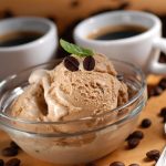 Gelato al caffè senza uova e senza gelatiera 1 Su tavolo chicchi di caffè sparsi. Primo piano di una ciotola di vetro con palline di gelato al caffè. Dietro una tazzina di caffè espresso e un cucchiaino