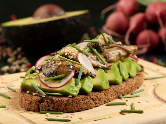 su tagliere di legno una fetta di pane integrale con sopra fettine di avocado, funghi porcini e ravanelli