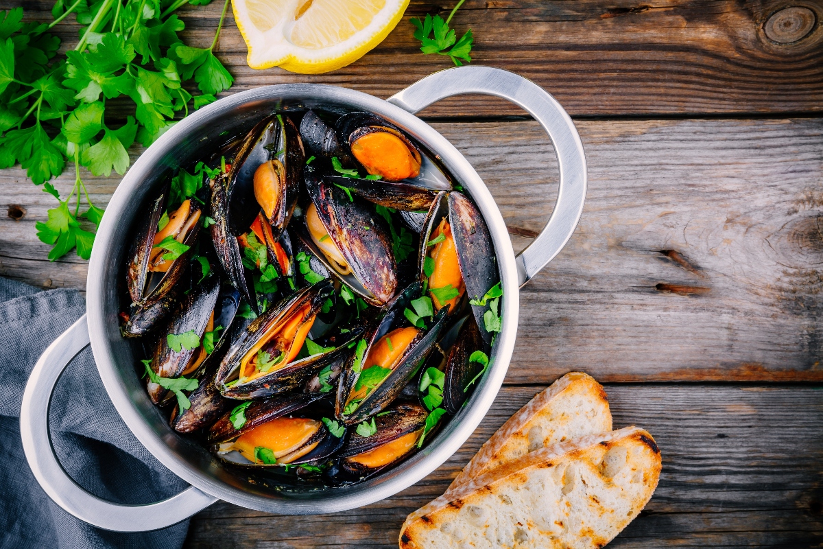 su tavolo di legno, padella per sauté con cozze alla marinara, attorno prezzemolo e 2 bruschette