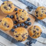 Muffin ai mirtilli con yogurt, ideali per colazione e merenda 1 In primo piano su una tovaglietta a righe bianca e blu e un tagliere di legno, ci sono dei muffin ai mirtilli con dei mirtilli freschi accanto.