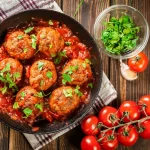 Polpette al sugo: come farle morbide e succose 1 padella tonda piena di polpette al sugo, ciotola di basilico e pomodorini accanto