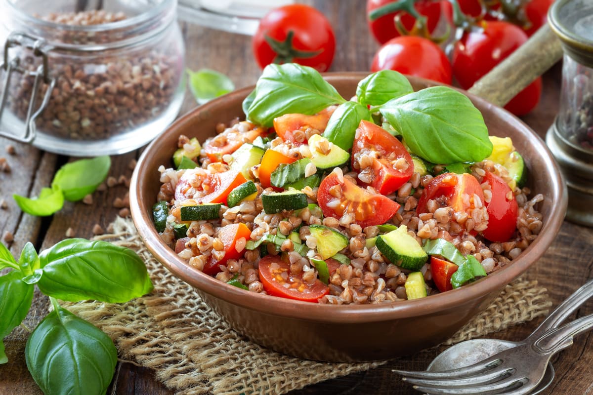 In primo piano su una tovaglietta di iuta c'è un piatto fondo con del farro , zucchine, pomodori e delle foglie di basilico. Sullo sfondo ci sono dei pomodori freschi e un barattolo di farro.