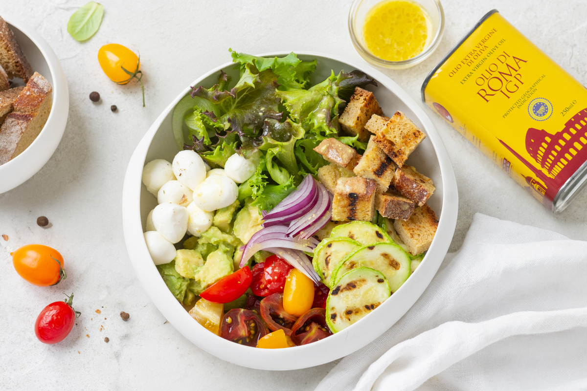Insalate sfiziose: le migliori ricette sane da provare a casa 14 In primo piano, su uno sfondo bianco, una ciotola di insalata mista con verdure croccanti, pomodorini e mozzarelline. Sulla destra una lattina dell'olio di Roma IGP.