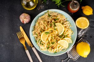 Iscriviti subito alla dieta Melarossa e scarica l'app 13 su tavolo di marmo nero piatto verde con pasta al limone una delle deliziose ricette con limone. Attorno limoni, una bottiglia di olio EVO, aglio e grattugia