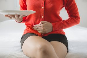 Iscriviti subito alla dieta Melarossa e scarica l'app 12 donna sul bordo del letto con cardigan rosso e shorts marrone, mano appoggiata sulla pancia e nell'altro mano un piatto bianco vuoto perché si chiede cosa mangiare con la diarrea