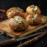 Patate al cartoccio: ricetta passo passo e video ricetta e come farle ripiene 1 su sfondo scuro tagliere di legno con 4 patate al cartoccio con ripieno di formaggio e prosciutto e erbe aromatiche , attorno coltelli e dietro specie