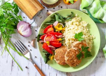 su tavolo bianco di legno un piatto verde con polpette di tonno, riso e peperoni. Attorno aglio, 1 forchetta e pepe macinato
