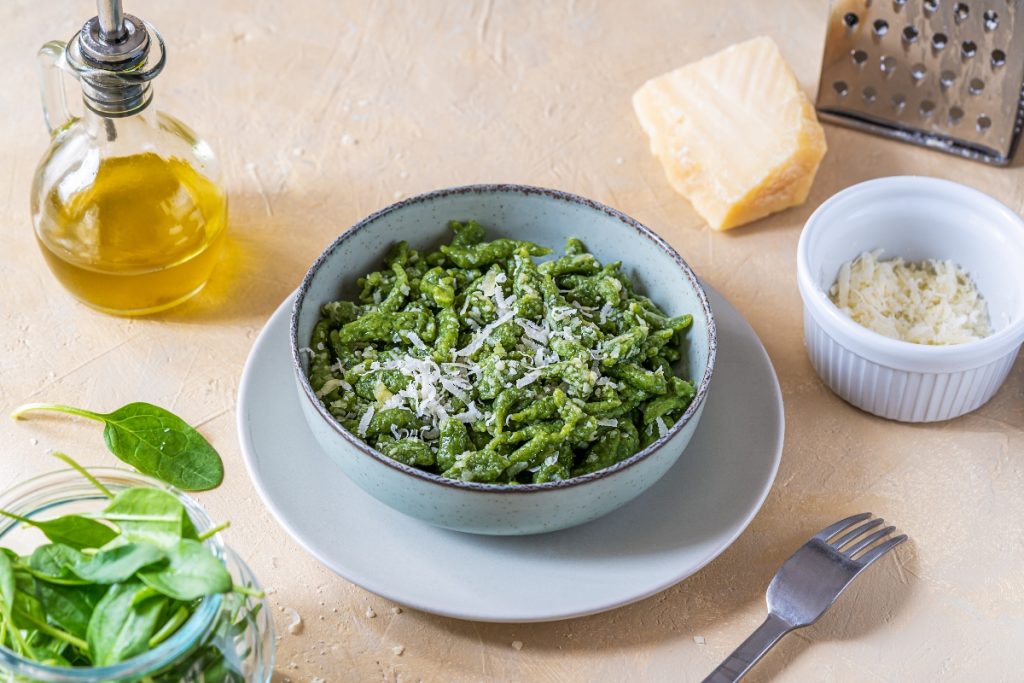 Spätzle: la guida completa all'originale ricetta tirolese 4 Su tavolo di legno chiaro ciotola con gli Spaetzle verdi fatti con gli spinaci, accanto spinaci freschi, una forchetta, olio EVO, parmigiano grattugiato