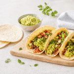 Tortillas di mais: la ricetta per farle a casa e come farcirle 1 Su un piano di marmo bianco c'è un tagliere di legno con delle tortillas alcune sono condite con avocado e dei fagioli. Sullo sfondo c'è una ciotola con della guacamole e un canovaccio bianco.