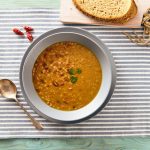 Zuppa di farro: la ricetta di un piatto unico nutriente e saziante 1 Zuppa di farro e fagioli servita in un piatto fondo su tovaglietta a righe, con pane su tagliere di legno su sfondo in legno chiaro.