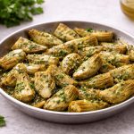 Carciofi trifolati in padella: un contorno versatile e vegetariano 1 In primo piano in un piatto fondo ci sono dei carciofi trifolati conditi con del prezzemolo e del peperoncino. Sullo sfondo degli spicchi d'aglio, olio evo e prezzemolo.