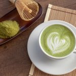 stuoia di bambù con piattino e tazza piena di matcha latte con un disegno a cuore, frusta e polvere matcha accanto