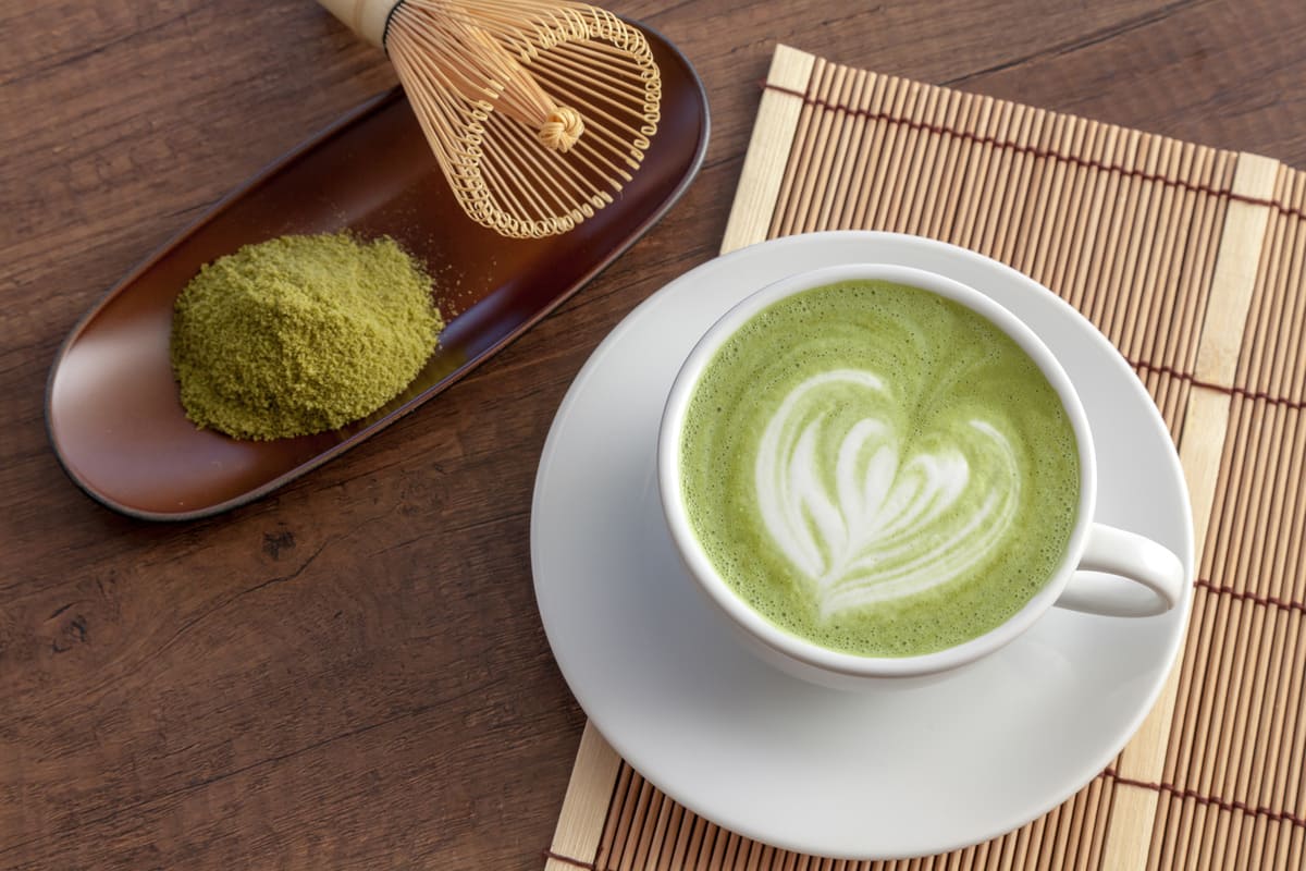 stuoia di bambù con piattino e tazza piena di matcha latte con un disegno a cuore, frusta e polvere matcha accanto