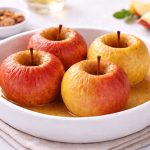 Mele al forno: ricetta facile per una merenda sana 1 In primo piano su una pirofila bianca ci sono delle mele al forno. Sullo sfondo ci sono dei spicchi di mela e una ciotolina bianca con della frutta secca.