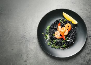 piatto nero su piano grigio con un risotto al nero di seppia, gamberi, prezzemolo e 1 fetta di limone