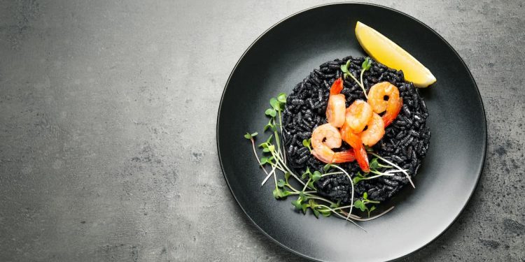 piatto nero su piano grigio con un risotto al nero di seppia, gamberi, prezzemolo e 1 fetta di limone