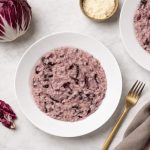 In primo piano su un tavolo di marmo chiaro c'è un piatto di risotto al radicchio. Sulla destra c'è una forchetta dorata con un tovagliolo color tortora. Sullo sfondo un cespo di radicchio rosso di Treviso.