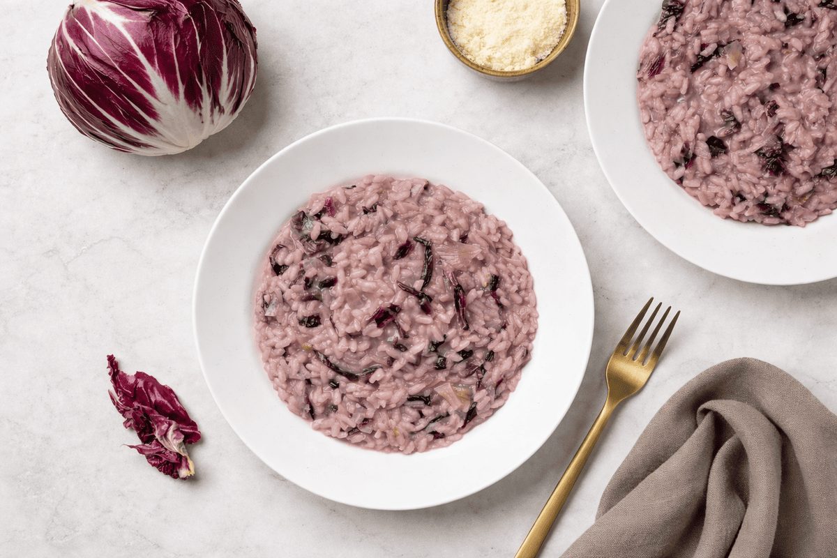 In primo piano su un tavolo di marmo chiaro c'è un piatto di risotto al radicchio. Sulla destra c'è una forchetta dorata con un tovagliolo color tortora. Sullo sfondo un cespo di radicchio rosso di Treviso.