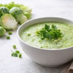 In primo piano su un tavolo di marmo bianco c'è un piatto con la zuppa di cavoletti di Bruxelles condita con delle foglie di prezzemolo. Sullo sfondo alla sinistra ci sono dei cavoletti di Bruxelles.