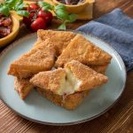 Mozzarella in carrozza: 1 In primo piano su un tavolo di legno c'è un piatto di mozzarella in carrozza, sullo sfondo delle ciotoline di legno con dei pomodori secchi.