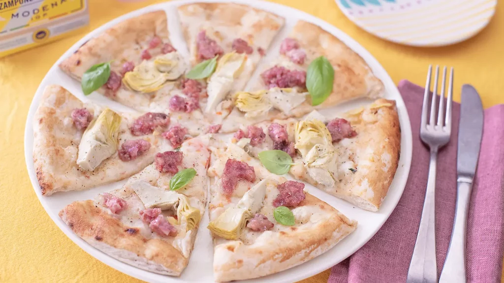 su un tavolo con tovaglia gialla, una pizza con cotechino e carciofi tagliata in 8 su un piatto bianco e accanto forchetta e coltello con un tovagliolo bordeaux