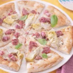 su un tavolo con tovaglia gialla, una pizza con cotechino e carciofi tagliata in 8 su un piatto bianco e accanto forchetta e coltello con un tovagliolo bordeaux