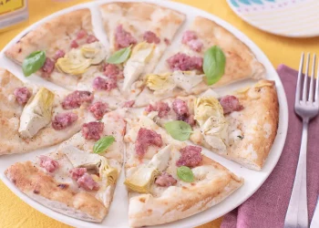 su un tavolo con tovaglia gialla, una pizza con cotechino e carciofi tagliata in 8 su un piatto bianco e accanto forchetta e coltello con un tovagliolo bordeaux