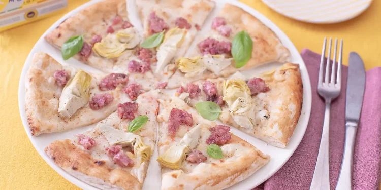 su un tavolo con tovaglia gialla, una pizza con cotechino e carciofi tagliata in 8 su un piatto bianco e accanto forchetta e coltello con un tovagliolo bordeaux