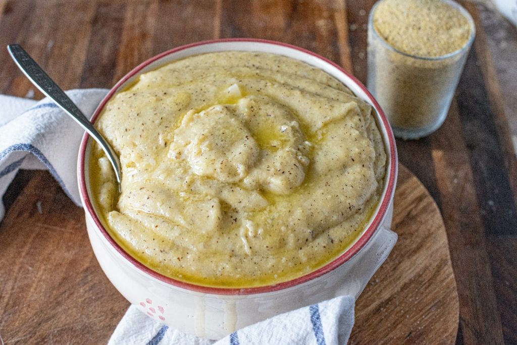 Polenta taragna: un piatto nutriente della tradizione valtellinese 3 su tavola di legno, ciotola con la polenta taragna e un cucchiaio, accanto un bicchiere di farina di grano saraceno. Sotto la ciotola, c'è un canovaccio bianco