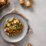 Topinambur in padella: contorno veloce con pochi ingredienti e tante varianti 1 In primo piano su un tavolo grigio, c'è un piatto di topinambur in padella con del prezzemolo sopra. Sullo sfondo ci sono delle radici di topinambur e dei macina sale e pepe.