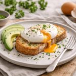 In primo piano su un tavolo di legno c'è un piatto con uova in camicia su una fetta di pane tostato, dell'erba cipollina tritata sopra e delle fettine di avocado.