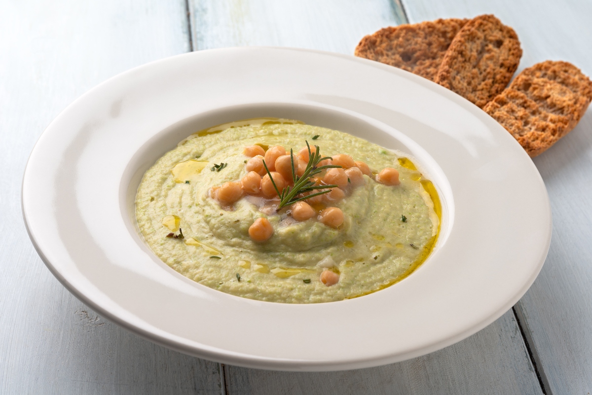 su tavolo di legno bianco, piatto bianco fondo con vellutata di ceci e alcuni ceci su vellutata accanto sulla destra 3 crostini