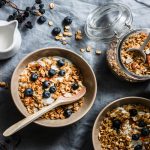 In primo piano su una tovaglia scura, due ciotole con della granola fatta in casa, dello yogurt e dei mirtilli. Sulla destra un barattolo in vetro che conserva della granola mentre sulla sinistra c'è un bricchetto con del latte.