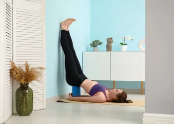 donna in casa che esegue u esercizio di pilates al muro contro una parete blu con un mattone Yoga dietro alla schiena e le gambe in alto