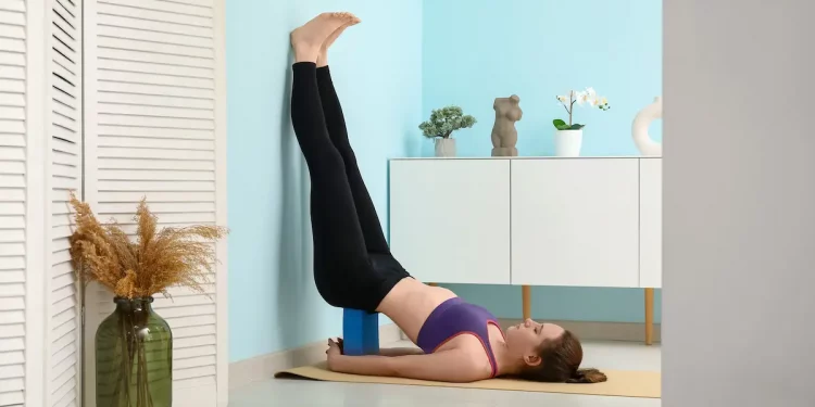 donna in casa che esegue u esercizio di pilates al muro contro una parete blu con un mattone Yoga dietro alla schiena e le gambe in alto