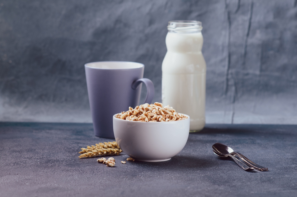 su sfondo grigio una tazza grigia con una bottiglia di vetro riempita di latte e una tazza di farro soffiato con spelta. Accanto 2 cucchiaini e sulla destra grani di frumento