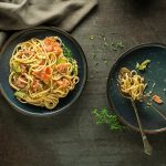 Pasta al salmone con ricotta: cremosa e senza panna 1 su tavolo di legno, 2 piatti neri con pasta al salmone. nel primo piatto sulla sinistra c'è tutta la pasta, mentre nell'altro piatto è quasi finito c'è forchetta e cucchiaio con spaghetti arrotolati . sulla sinistra bicchiere di acqua con 1 fetta di limone.