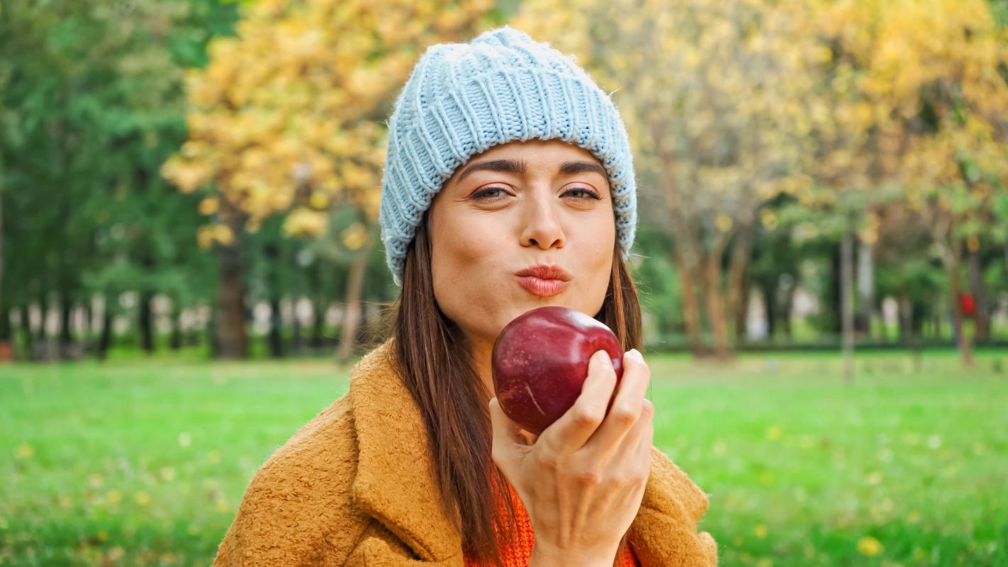 Bellissima donna con capotto e capello al parco guarda drittol'obbiettivo sorridendo e mangiando una mela rossa